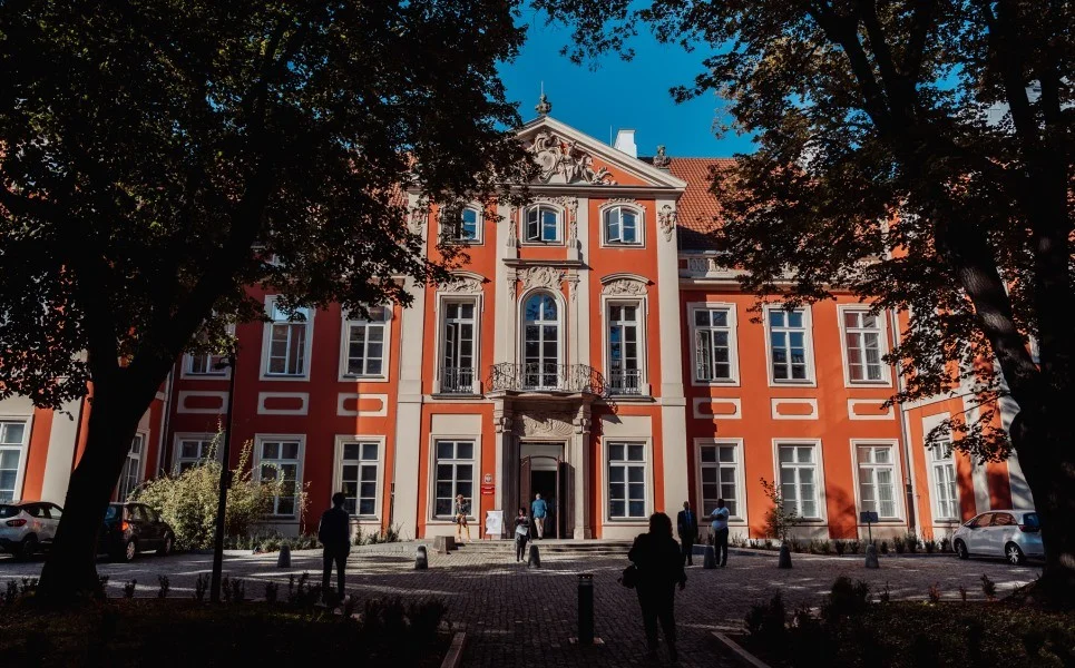 Kremowo pomarańczowy zabytkowy budynek Akademii Sztuk Pięknych w Warszawie. Front wyeksponowany, zdobiony. Boki budynku przysłaniają drzewa