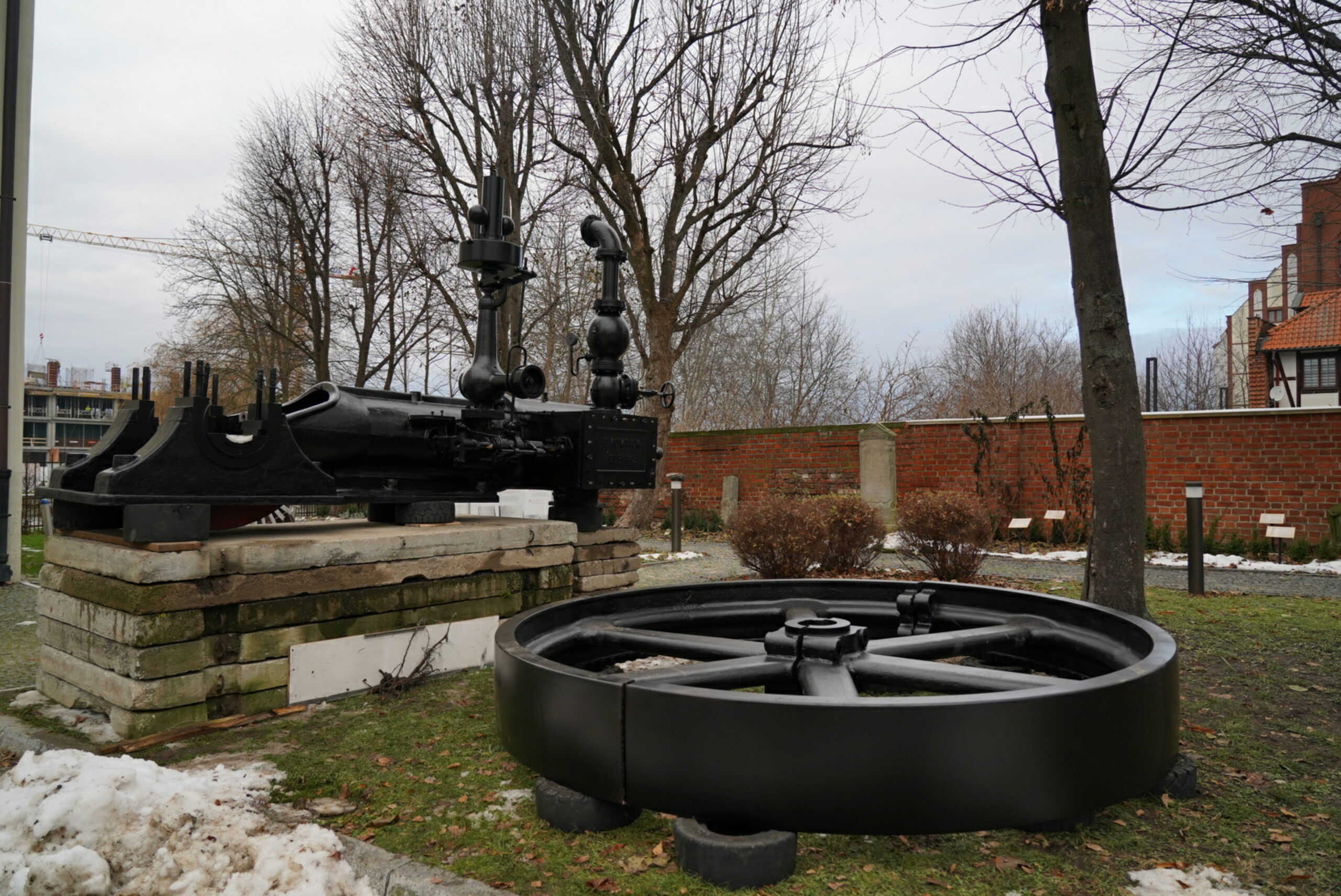 Zdjęcie przedstawia maszynę parową stojącą na kilku położonych jedna na drugiej płytach betonowych. Obok na oponach położono masywne, duże stalowe koło. Wszystko pomalowane na czarno.