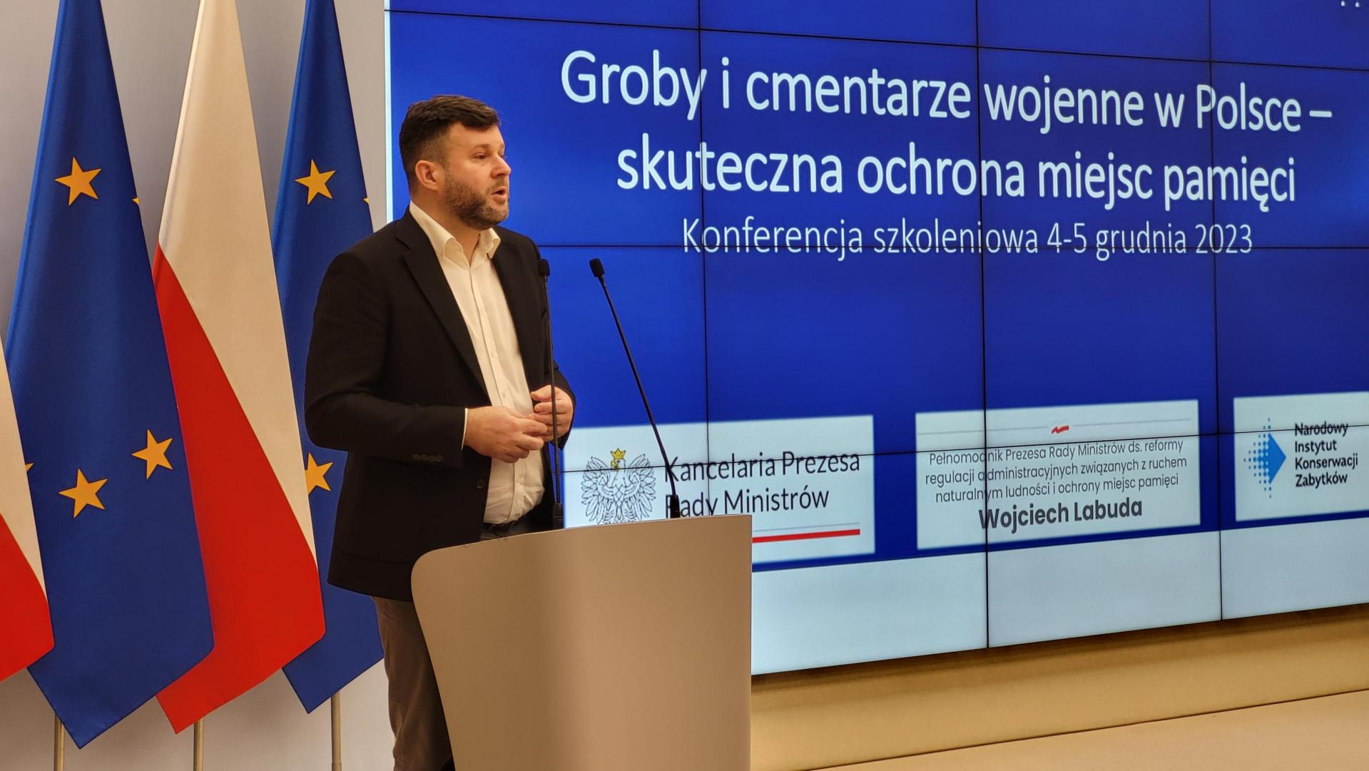Prelegent na scenie omawiający prezentacje. W tle flagi Polski i UE i ekran z prezentacją