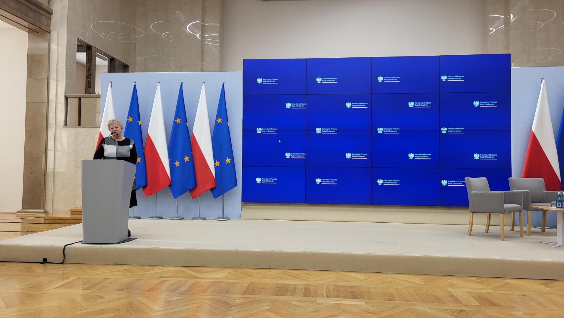 Scena z mównicą oraz w tle flagi Polski i UE. Obok duży ekran do prezentacji
