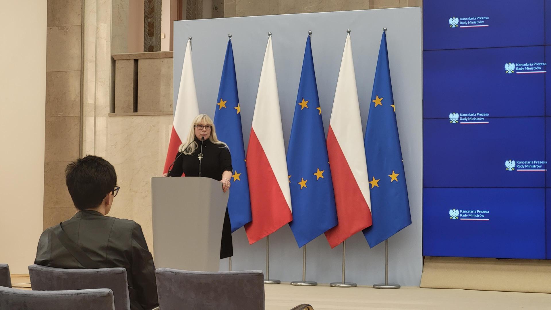 Scena z mównicą przy której stoi Dr. Agnieszka Partridge, w tle flagi Polski i UE. Obok duży ekran do prezentacji