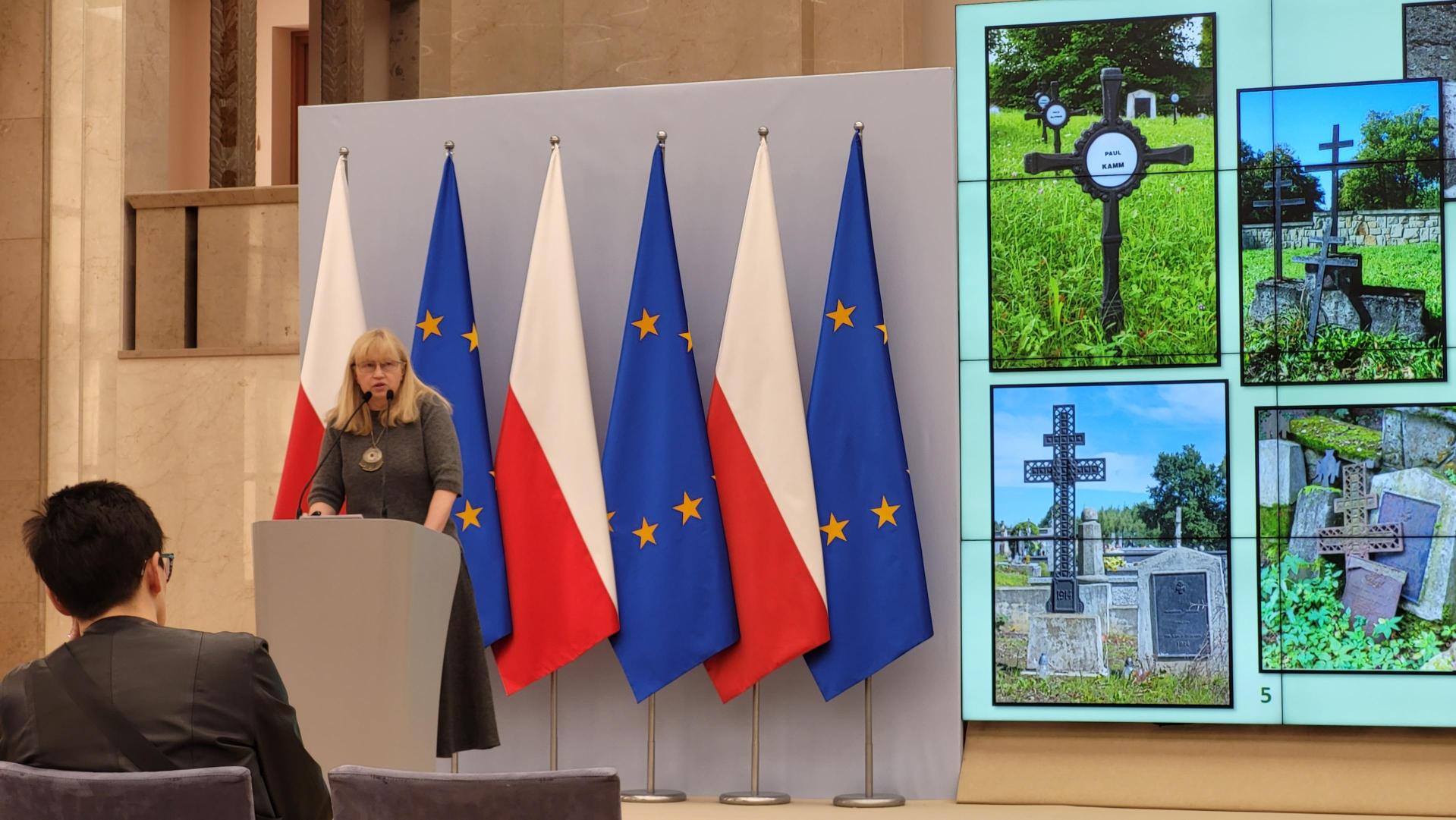 Scena z mównicą przy której stoi Dr. Beata Nykiel, w tle flagi Polski i UE. Obok duży ekran do prezentacji